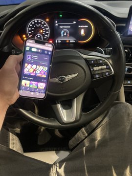 A photo showing a hand holding a smartphone displaying a purple-themed app interface in front of a Genesis car steering wheel, with the vehicle's digital dashboard illuminated in the background.