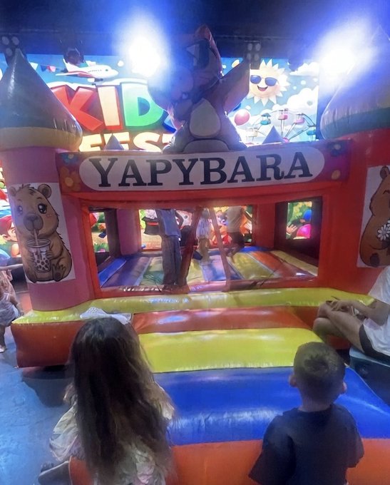 Children watch an inflatable bouncy castle attraction called 'YAPYBARA' at what appears to be a 'Kids Fest' event, featuring cartoon animal illustrations on the castle towers.
