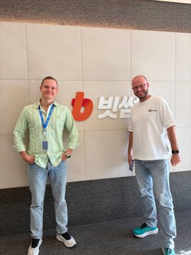 Two men pose for a photo in an office setting in front of a wall with the '비상' (Bisan) company logo.