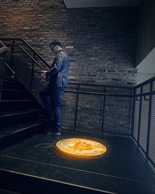 A man in a blue jacket stands on dark stairs in an industrial brick-walled space, while a glowing Bitcoin projection illuminates the floor below.