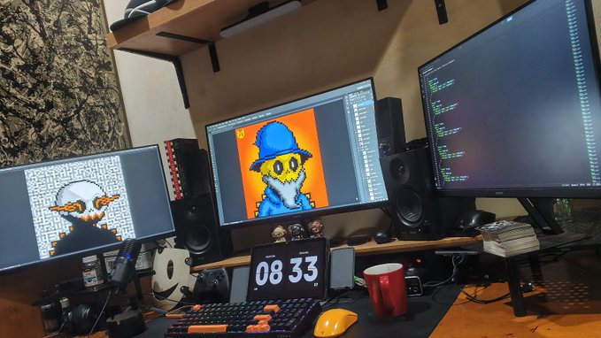 A desk setup with multiple monitors displaying pixel art characters, a digital clock reading 08:33, speakers, a keyboard, and various desk accessories.