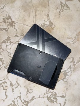 A sleek black metal X (formerly Twitter) branded card from X Money lies on a marble surface, featuring a minimalist design with the X logo and "X MONEY" text engraved on its front. The card represents the user's new direct deposit setup for the X Money platform, which they're excited to potentially use for crypto purchases in the future.