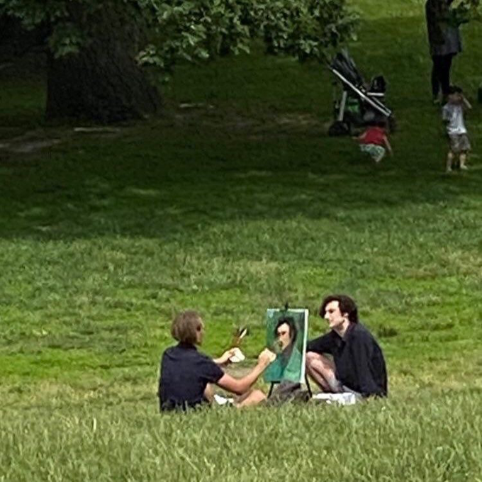 A candid outdoor scene in a park where a person is sitting on the grass painting a portrait of another person seated next to them, with a stroller and other park-goers visible in the background.