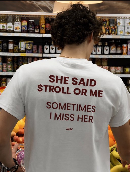 A man wearing a white t-shirt with the text 'SHE SAID $TROLL OR ME SOMETIMES I MISS HER' stands in front of a grocery store shelf stocked with various products.