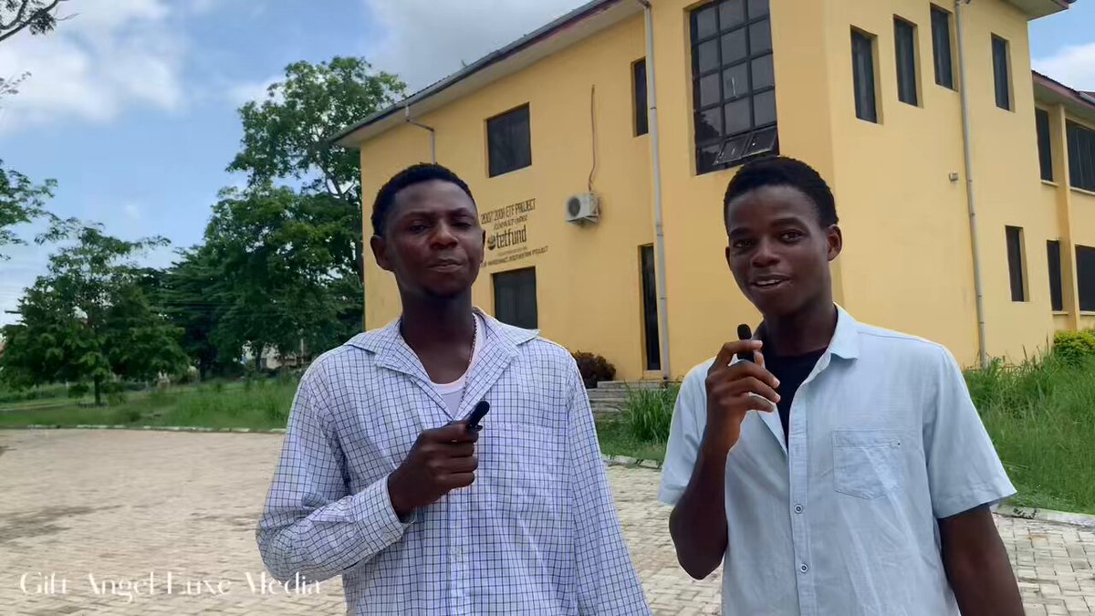 The image shows two young men standing outside, each holding a cell phone. They appear to be posing for a photo or recording a video related to Bitcoin Pizza Day. The background features a yellow building with windows, which could possibly be an office or a store associated with Bitcoin Pizza Day.