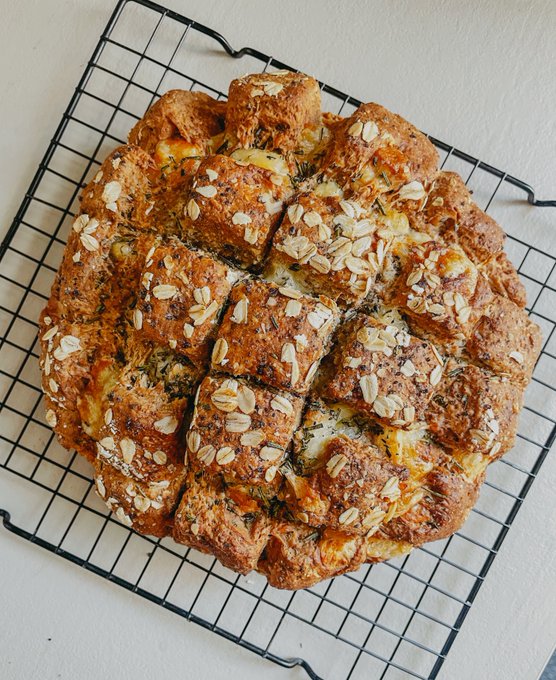 A freshly baked round loaf of bread, topped with melted cheese, rolled oats, and herbs, sits cooling on a black wire rack, illustrating the "cheese soda bread" referenced in the post by the #cryptosoupgroup.