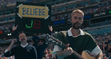 A man plays acoustic guitar while holding a 'BELIEBE 4:43' sign in a stadium filled with fans, referencing a Justin Bieber tribute or countdown.
