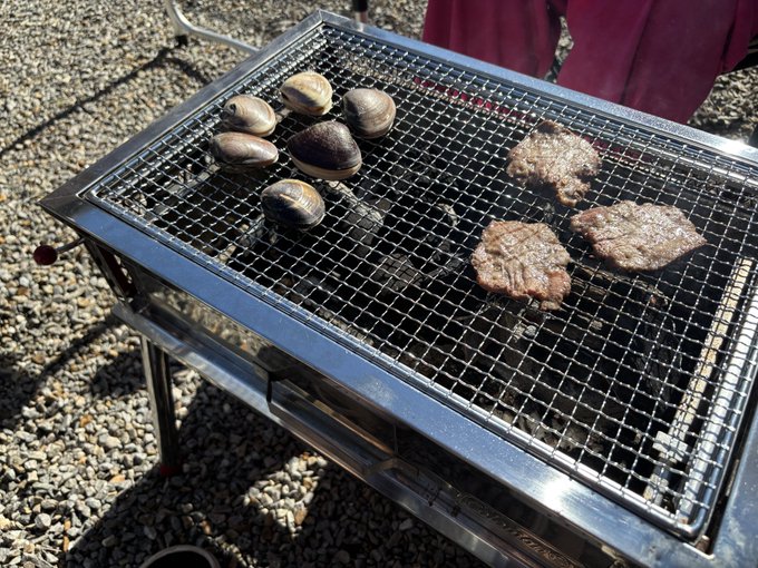 A portable metal grill sits on gravel, cooking several clams in their shells alongside grilling hamburger patties with visible char marks.