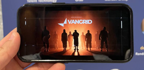 A hand holds a smartphone displaying the game VANGRID, which shows silhouetted figures standing against an orange, smoky background.