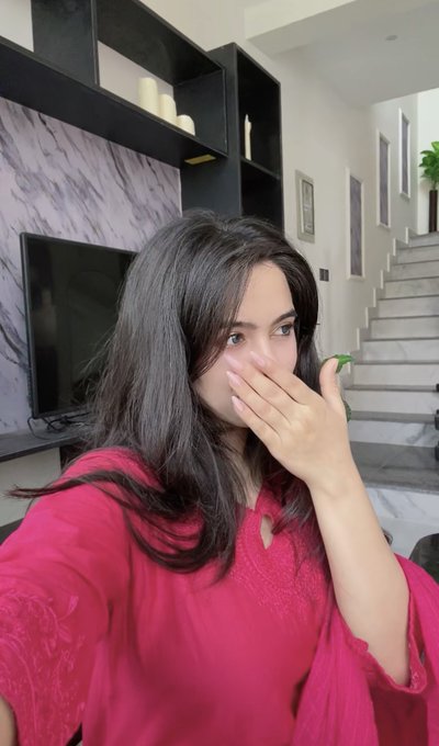 A young woman with long dark hair wearing a pink top covers her mouth with her hand while taking a selfie in a modern home interior.