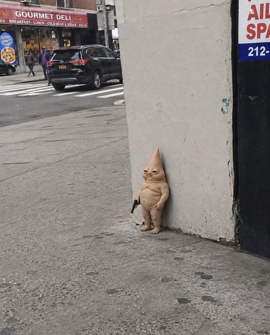 A small, gnome-like figurine with a pointed head and stern expression stands against a concrete wall on a New York City street, holding what appears to be a small object, with a "GOURMET DELI" sign and the partial numbers "212-" visible in the background. The post's playful, meme-like tone about a "next 100x runner" is humorously juxtaposed with the quirky, randomly-placed street ornament that has seemingly been "called upon" like a financial oracle.