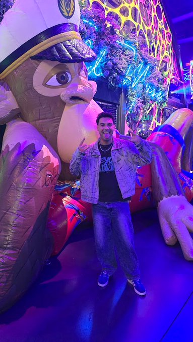 A man in a denim jacket poses in front of a large, illuminated mascot statue, possibly at an arcade or themed venue, with vibrant neon lighting in the background.