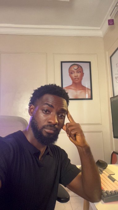 A bearded man sits thoughtfully with his finger to his temple in front of a computer setup, while a framed portrait of a woman with a forehead jewel hangs on the wall behind him, capturing the one-man production struggle of filming content alone.