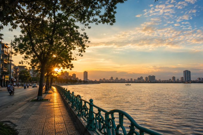 A scenic sunset view along a waterfront promenade with trees, a turquoise railing, a road with motorbikes, and a city skyline across the river.