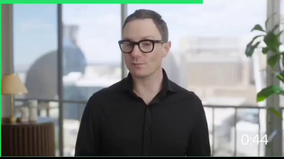 The image features a man wearing glasses standing in front of a window with a city skyline visible outside. The man appears to be smiling at the camera, possibly posing for a photo or video. The background includes a lamp and a potted plant, adding to the urban atmosphere. The image also contains text that reads "BIG! $RippleX just dropped a fresh “ XRP in One Minute" with Hugo Philion explaining how Ripple is used as collateral through FXRP for lending, stablecoin borrowing, DeFi yield, and vaults on XRPL."