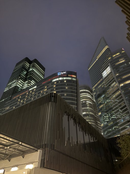 A nighttime view of modern skyscrapers, including one with a "lyf" sign, viewed from below against a purple-blue sky.