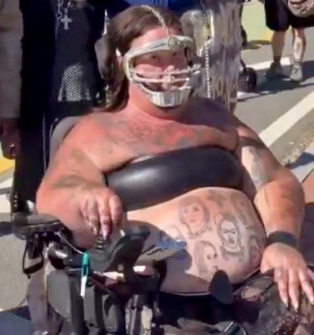 A heavily tattooed woman wearing a metallic face mask sits in a wheelchair, appearing at what looks like an outdoor event or festival. The image humorously illustrates the post's point about Bitcoin's "spam" problem by visually representing overwhelming, unwanted content—likely referencing the proliferation of BRC-20 tokens, Ordinals, or meme coins cluttering the Bitcoin network with low-value transactions.