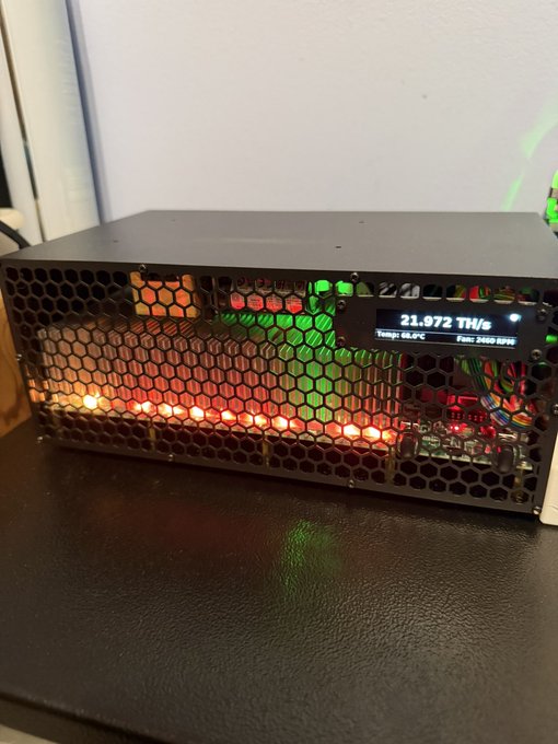 A GekkoScience KBOX home Bitcoin miner (serial #17) is illuminated with colorful LED lights through its honeycomb mesh exterior, with its display reading "21.972 TH/s" at a temperature of 53.0°C and fan speed of 2580 RPM. The device appears to be a compact, open-frame mining unit sitting on a dark surface, showcasing its impressive hash rate performance as mentioned in the post.