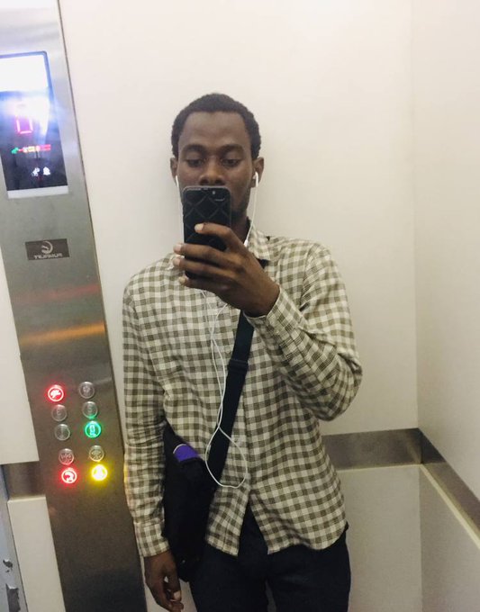 A young man wearing a green checkered shirt takes a mirror selfie in an elevator while listening to music through white earbuds connected to his smartphone, with the Hyundai elevator control panel visible behind him displaying illuminated floor buttons.