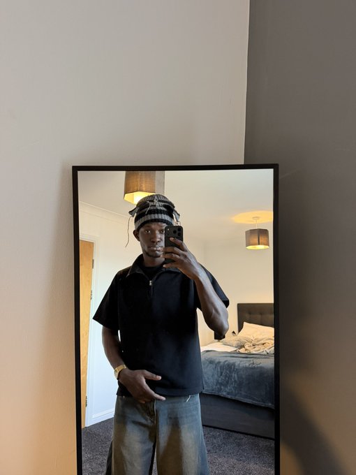A mirror selfie of a young Black man wearing a black short-sleeve shirt, gray jeans, and a patterned bandana, taking a photo in a hotel room or apartment.