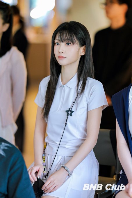 A young woman with long dark hair wearing a white polo shirt with a star logo sits in a crowd, with 'BNB Club' watermarked in the corner.