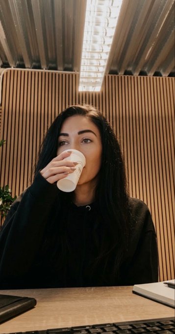 A young woman with long dark hair takes a sip from a white cup while sitting at a desk with a keyboard in the foreground.