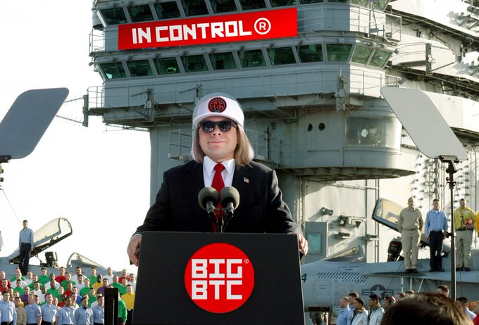 A man in a dark suit, red tie, white captain's hat, and sunglasses speaking at a podium on what appears to be an aircraft carrier, with a red 'IN CONTROL ®' sign above the ship's tower and a 'BIG BTC' logo on the podium.