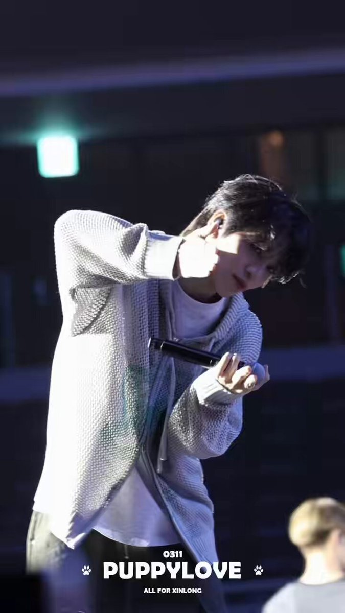 The image features a man wearing a white sweater, standing on stage with his hand up to his face as if he's singing or speaking into a microphone. The background of the photo shows a building, which could be part of an event taking place at the venue where the performance is happening. There are also some texts visible in the image that read "260613 #XINLONG #鑫隆 허씬롱 #ALPHADRIVEONE #シンロン #ホーシンロン".