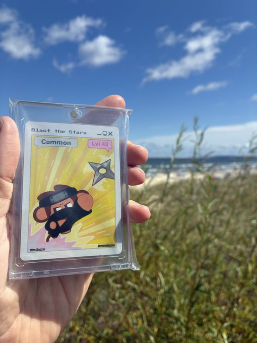A hand holding a graded trading card titled 'Blast the Stars' (Common, Lvl 42) featuring a cartoon character, set against a blue sky and grassy outdoor background.