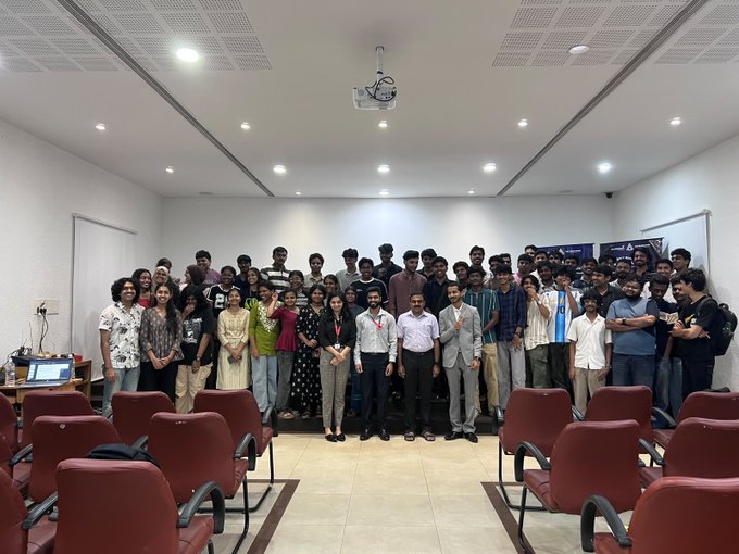 A group photograph of approximately fifty young adults and a few older individuals posing together in a modern auditorium or conference room with red seating.