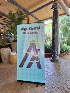 The image features a large blue banner with an "A" on it, standing upright against a brick wall or stone floor. The banner displays the text "AlgoBharat Hack Series", indicating that this event is related to AlgoBharat and involves a hackathon. There are also potted plants in the background, adding some greenery to the scene.