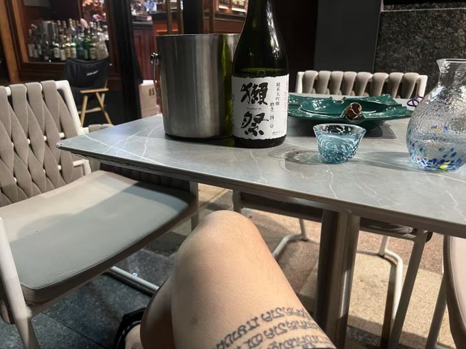 A first-person view of a person's leg with a tattoo, sitting at a marble cafe table holding a sake bottle (禪, 'Dassai'), a metal ice bucket, and small glass cups.