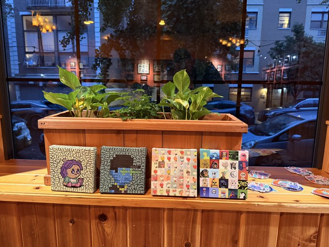 A wooden planter box with green leafy plants sits on a counter behind small decorative tile artworks, with a window showing a city street and buildings at dusk in the background.