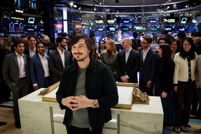 A man stands smiling in front of a podium with a decorative frame, surrounded by a crowd on the floor of the New York Stock Exchange, where the "HOOD" ticker for Robinhood is displayed on screens in the background.
