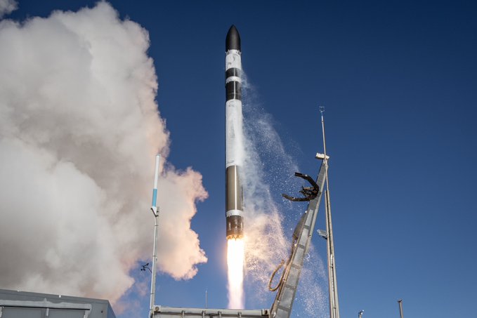 A Rocket Lab Electron rocket lifts off from its launch pad against a clear blue sky, engulfed in bright orange flames and billowing white exhaust clouds as it ascends vertically past the launch tower structure, capturing the triumphant moment of a successful mission launch on a bright, sunny day.