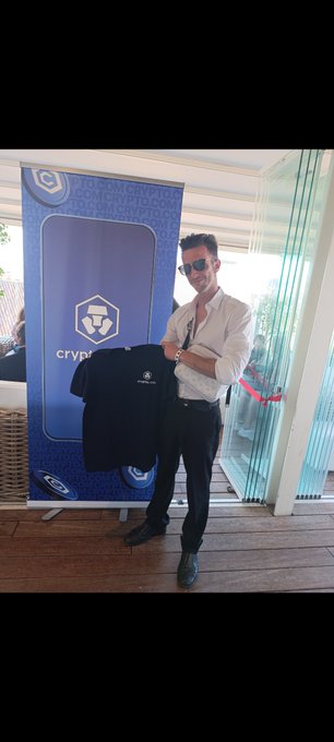A man in sunglasses and a white shirt stands in front of a Crypto.com banner, holding up a black t-shirt at what appears to be a promotional event.