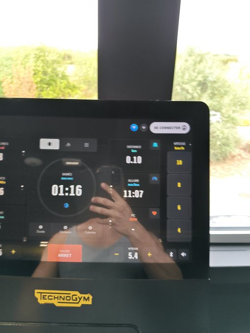 A person's reflection is visible on the screen of a TechnoGym exercise machine displaying a workout timer of 01:16, with a window showing greenery in the background.