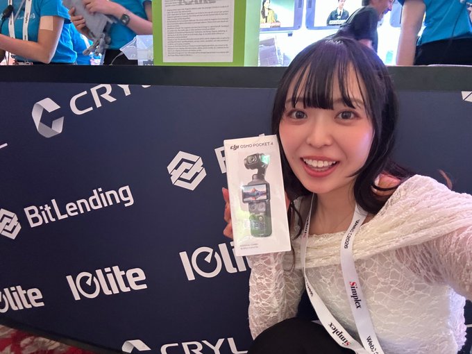 A smiling young woman poses at a trade show booth with 'BitLending', 'Iolite', and 'CRYPTO' logos, holding a box for a 'DJI OSMO POCKET 4' camera.