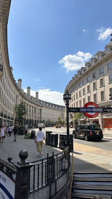 A sunny street view of London's Piccadilly Circus area, featuring classic curved architecture, a black taxi, pedestrians, and a London Underground sign for the Piccadilly line.