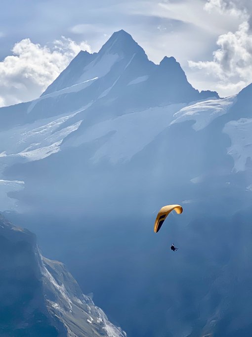 A paraglider with an orange canopy soars through misty air beneath towering snow-capped mountain peaks beneath a partly cloudy sky.