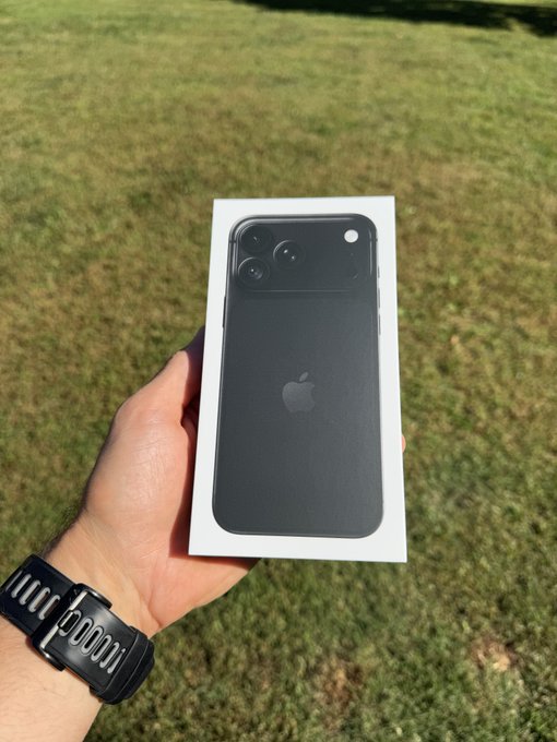 A hand wearing a black smartwatch holds a white box containing a black (likely graphite) iPhone outdoors over grass.