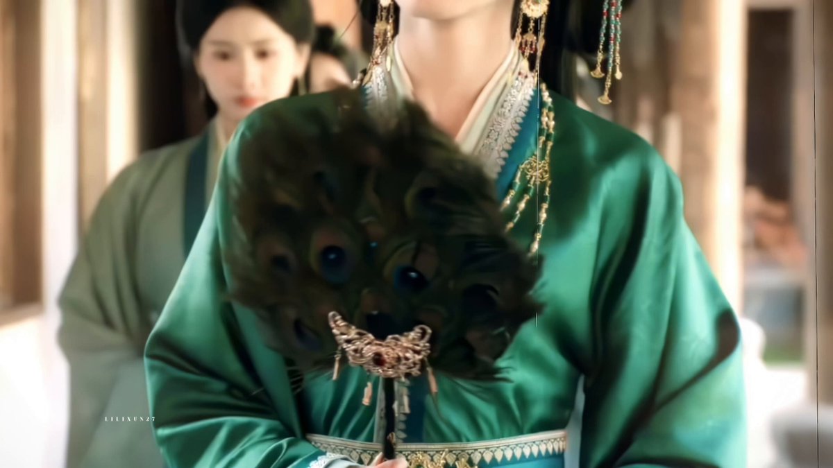 The image features a woman wearing a green dress with a peacock feather headpiece. She holds a large fan in her hands while standing next to another person who is also dressed in a green outfit, possibly a traditional Chinese outfit. The background of the image shows a hallway or room setting, which adds context to the scene and suggests that they might be in an area where people gather or socialize.