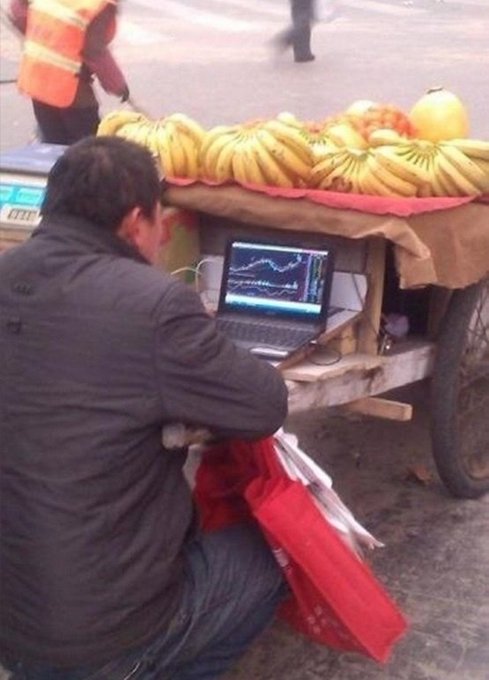 A fruit vendor is intensely focused on his laptop screen displaying a stock trading chart, with bunches of bananas and other fruits displayed on his cart behind him, humorously illustrating the meme's caption about being completely absorbed ("locked in") in trading while supposedly working.