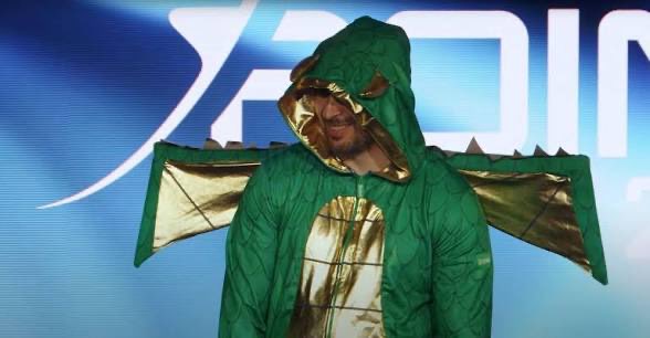 A person in a green and gold dragon costume stands in front of a blue screen displaying partial "KADIN" text. The costume features a hooded head, spiked back crest, and outstretched wings, resembling a dragon mascot outfit.