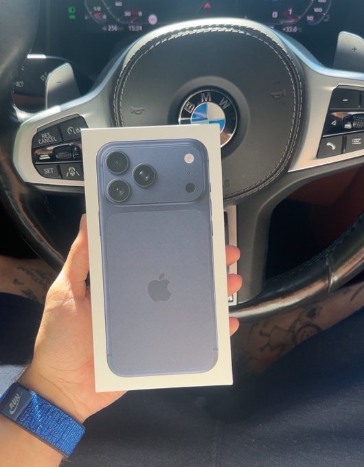 A person's hand holds a boxed blue iPhone Pro Max in front of the steering wheel of a BMW car, with a blue wristband visible on their wrist.