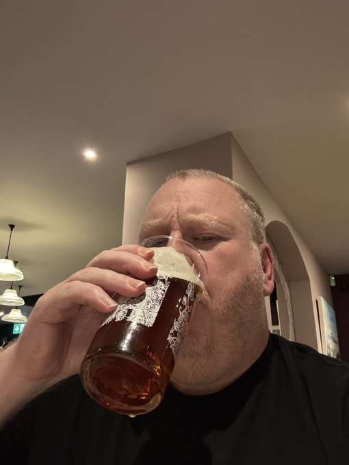 A close-up selfie of a bearded man drinking from a glass of beer in what appears to be a pub or restaurant setting.