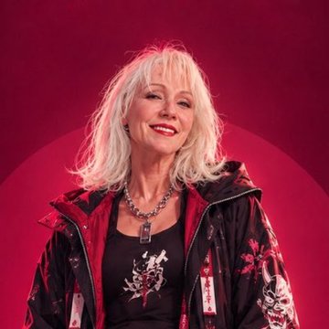 A smiling woman with voluminous white hair poses against a bright red background, wearing a red and black patterned jacket with a silver pendant necklace.