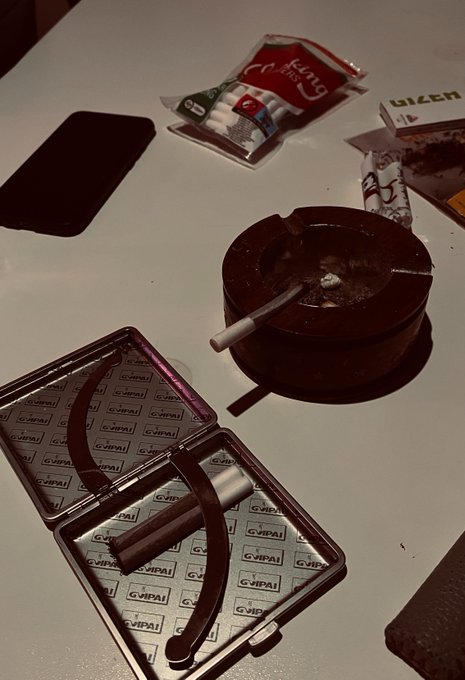 A dimly lit photo of a white table featuring a black smartphone, packs of chewing gum (Gang brand and Lizer), an open metal case labeled 'GIPAI', and a dark ashtray with a lit cigarette.