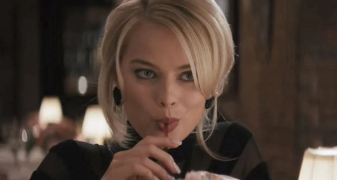 A scene from a film showing a blonde woman in a black top in a dimly lit restaurant, holding her hand near her mouth while drinking from a straw with an intense expression.