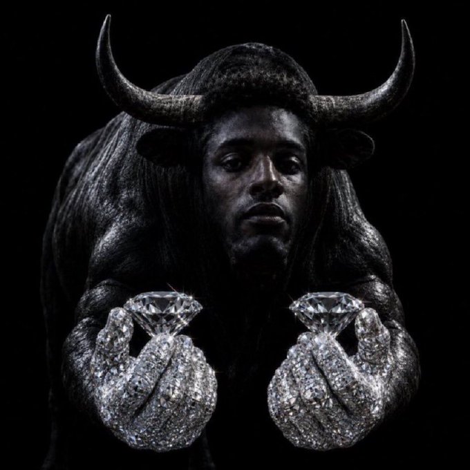 A dramatic, dark portrait shows a figure with bull horns and dark skin holding two large, sparkling diamonds in jewel-encrusted hands against a black background.